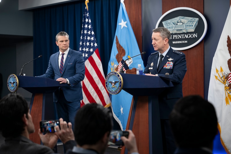 A man in business attire and a man in a formal military uniform stand behind lecterns and speak to the crowd before them. Behind them are an American flag, two flags with eagles and a sign that reads, The Pentagon, Washington.