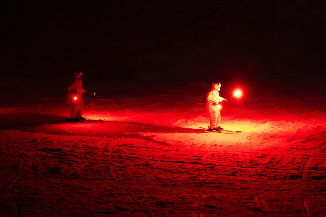 Two soldiers in ski gear carry red flares in the dark.