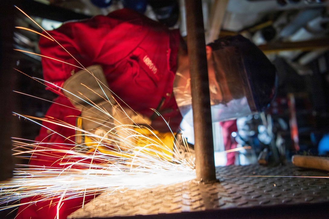 Sparks fly as a sailor in protective gear welds metal in a maintenance technician shop.