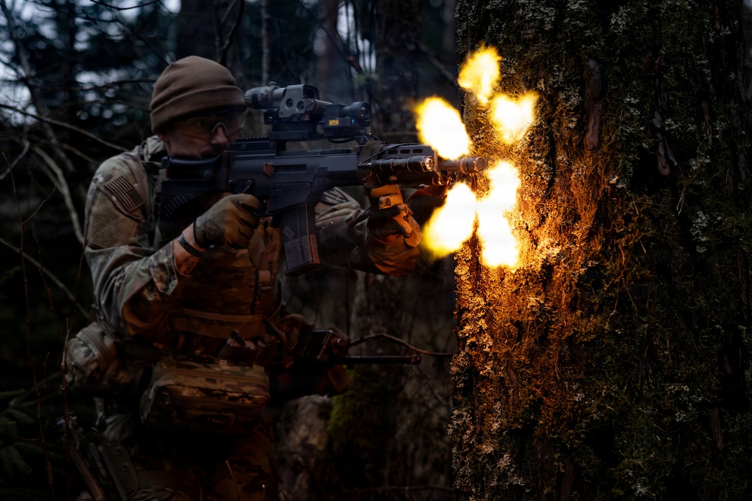 A soldier bends next to a tree in the dark to fire a weapon, creating orange fireballs.
