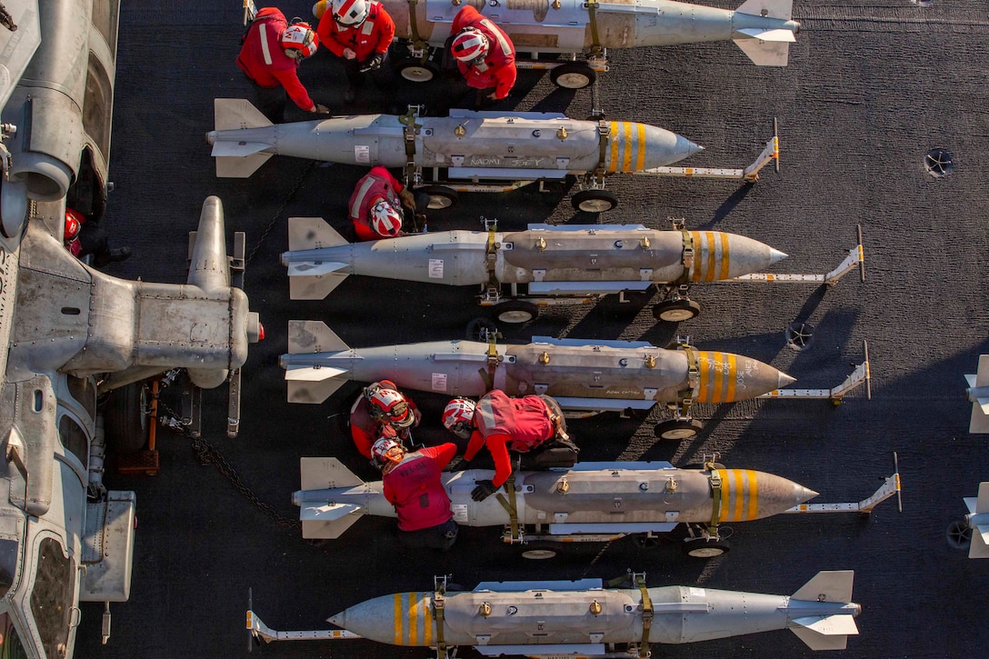 Sailors in red shirts and helmets work on ordnance aboard a ship as seen from above.