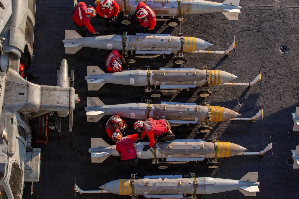 Sailors in red shirts and helmets work on ordnance aboard a ship as seen from above.