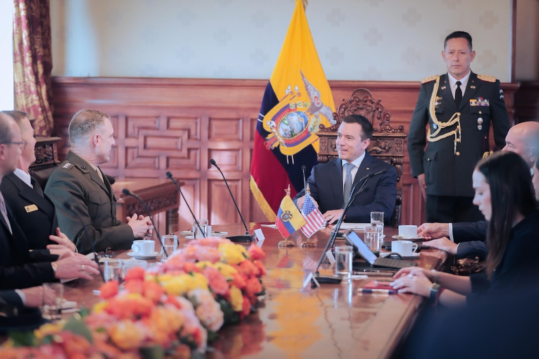 Senior officials in military uniforms and suits sit at a table while a man in a military uniform speaks with a man in a blue suit.