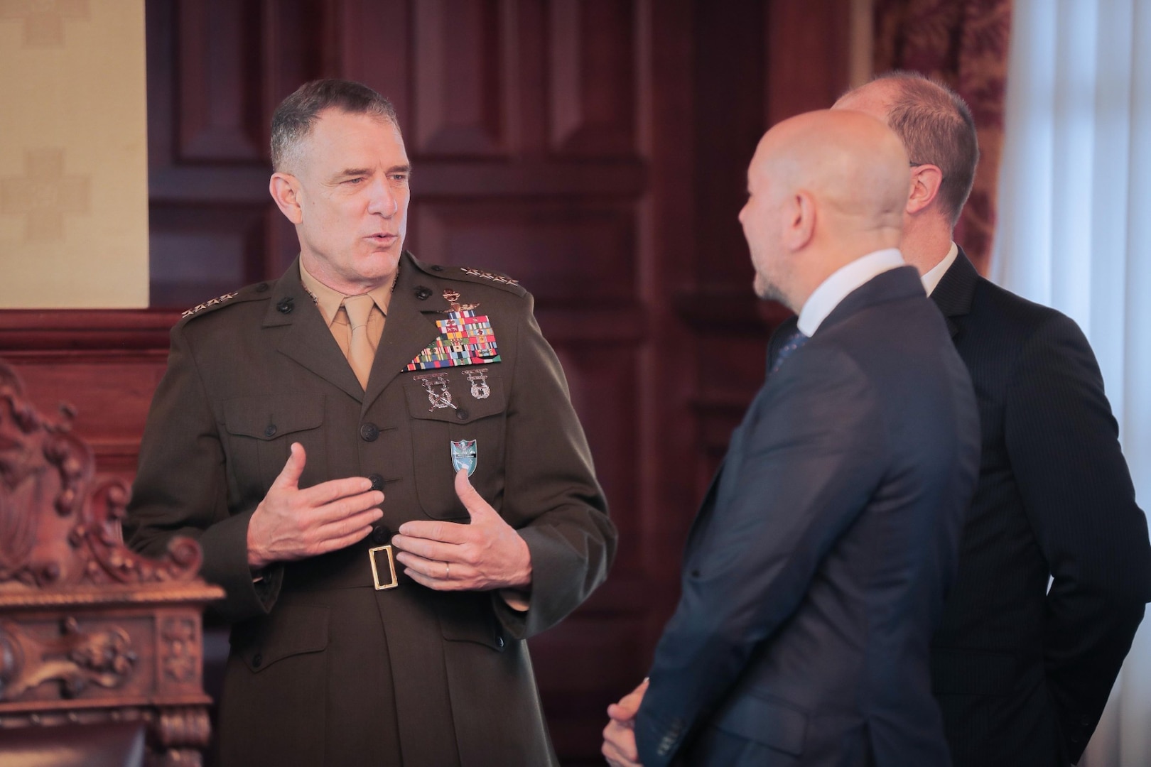 A man in a brown military dress uniform talks with a man in a blue suit.