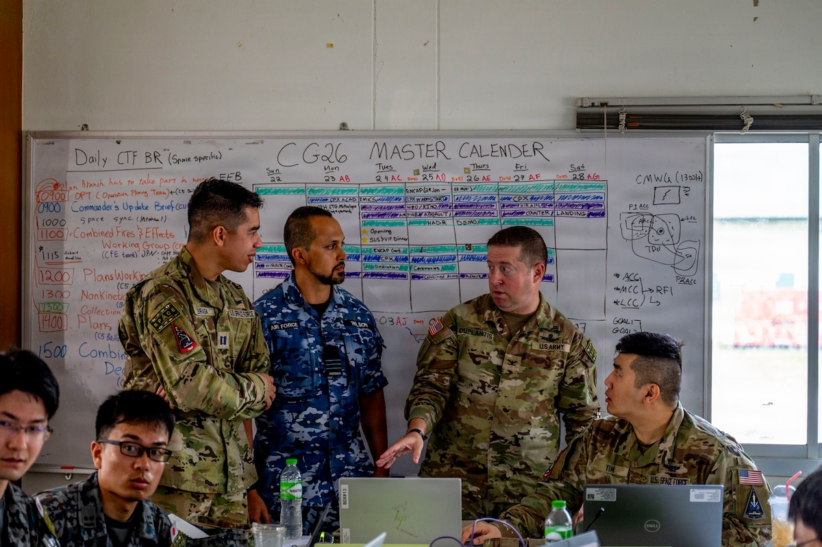 Three men wearing camouflage military uniforms from multiple nations stand in front of a white board. There are other men in similar attire seated at tables with laptops in front of them.