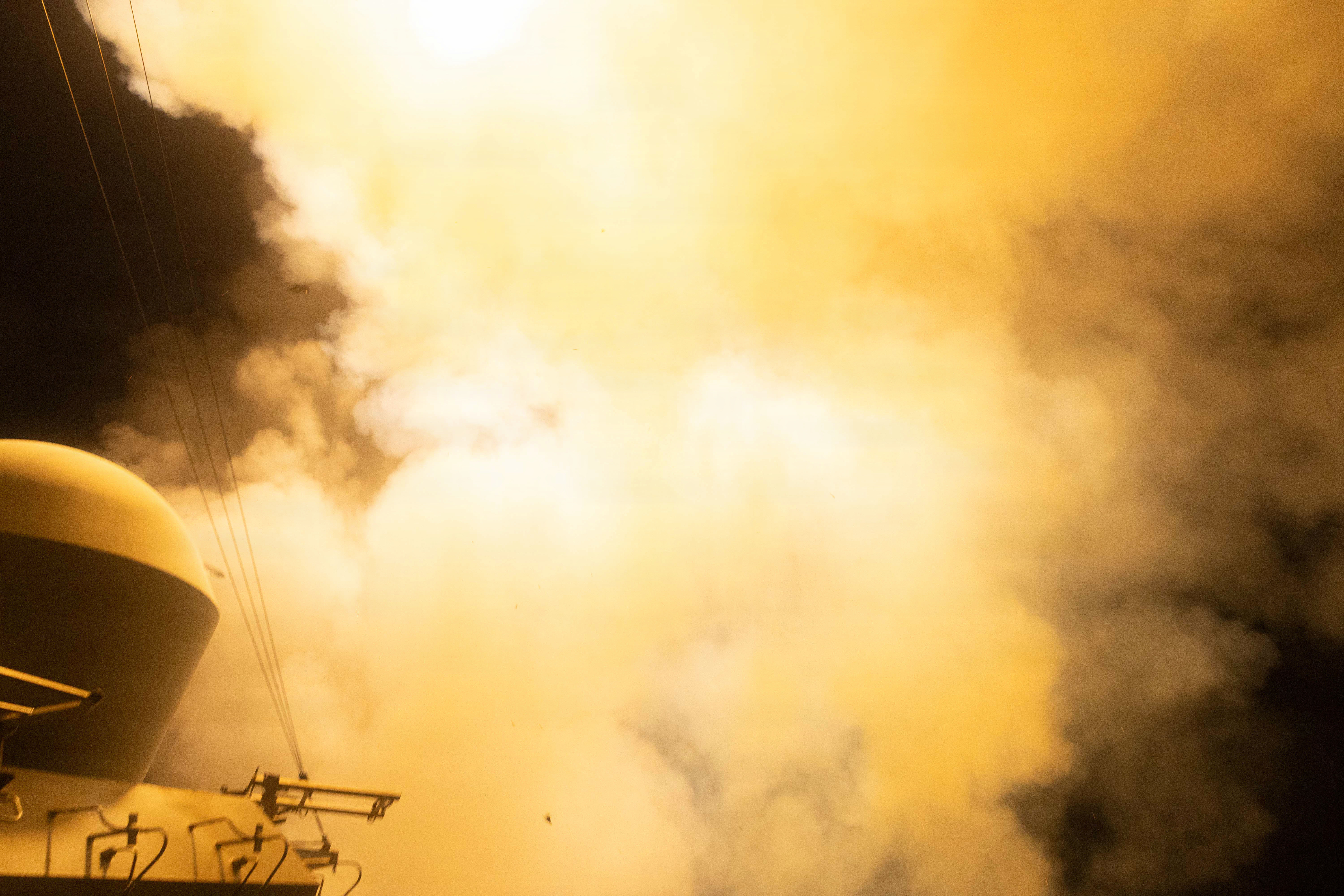 A missile launches from the deck of a ship in dark conditions, leaving orange flames and white smoke billowing.