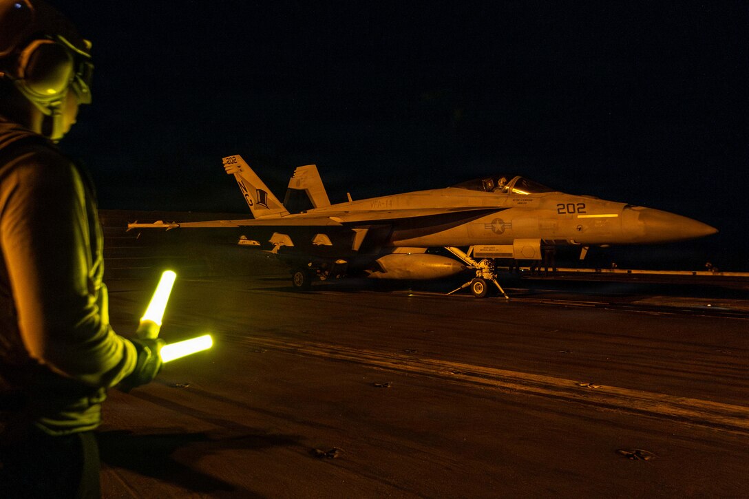 A fighter jet is seen on the deck of a ship in dark conditions, with a sailor nearby holding flashlights in each hand.