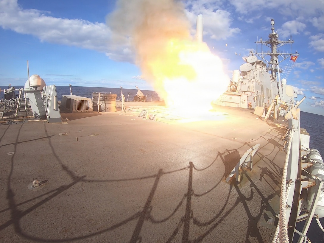 A missile launches from the deck of a ship during daytime, leaving flames and billowing smoke in its wake.