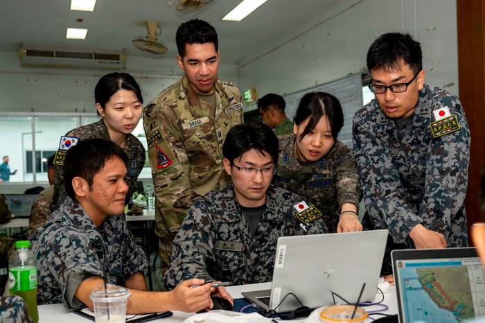 A group of people, both men and women and wearing various camouflage uniforms from multiple nations, stand and crouch as they look at a laptop on a table.
