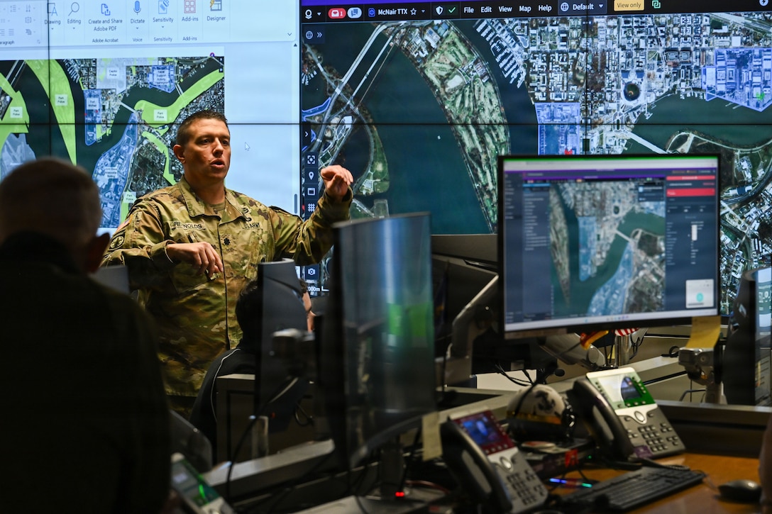 A soldier stands and talks in a dark room with maps on screens in the background and monitors in the foreground.