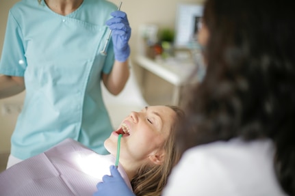 Dental patient wearing a pink bib opens her mouth as a dentist examines. The dentist's assistant stands behind the patient.