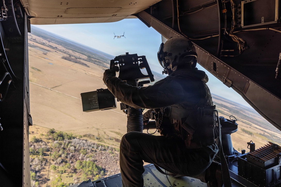 A Marine in tactical gear sits near an open doorway of an airborne aircraft as another aircraft flies in the distance over a desert-like area.