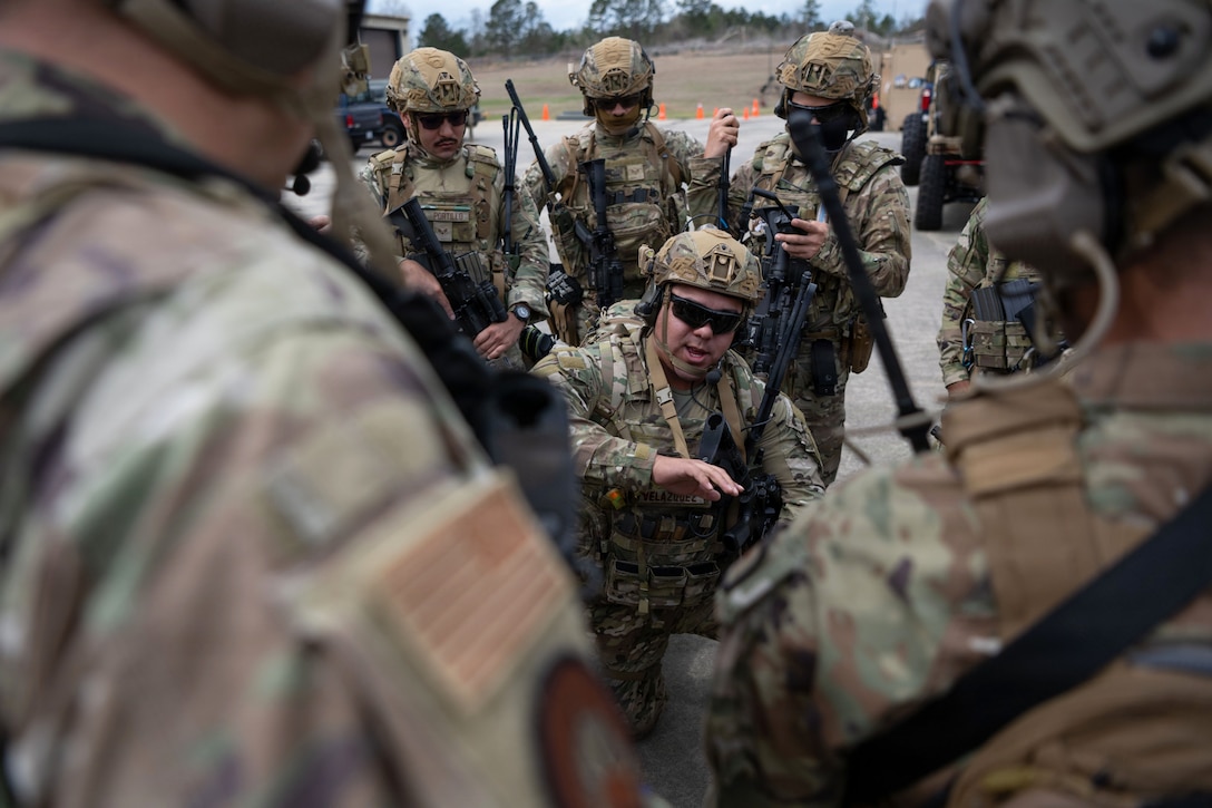 Airmen in tactical gear huddle around a kneeling airman in a parking lot near a field.