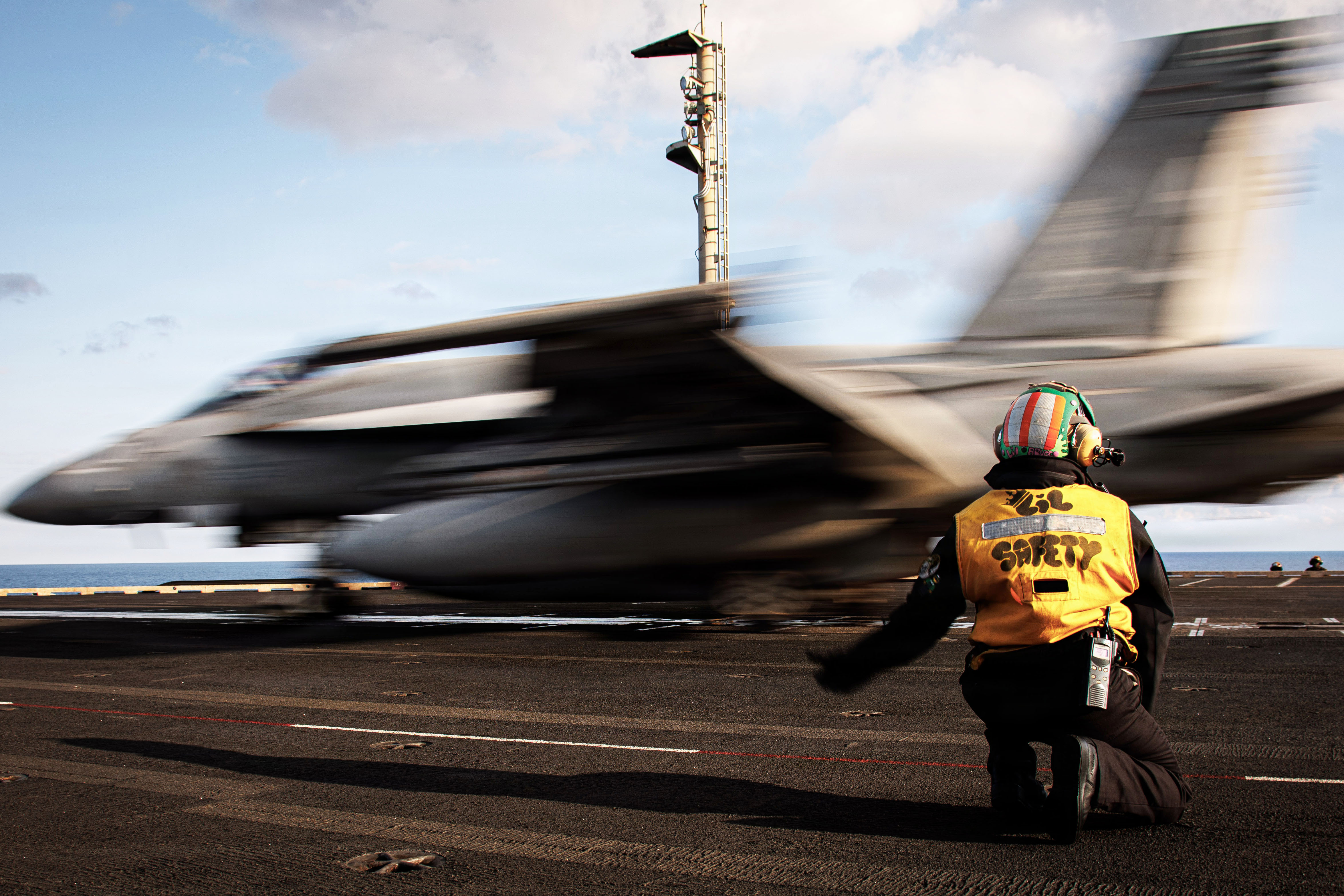 A kneeling sailor signals to a blurry aircraft as it launches from a ship at sea during daytime.