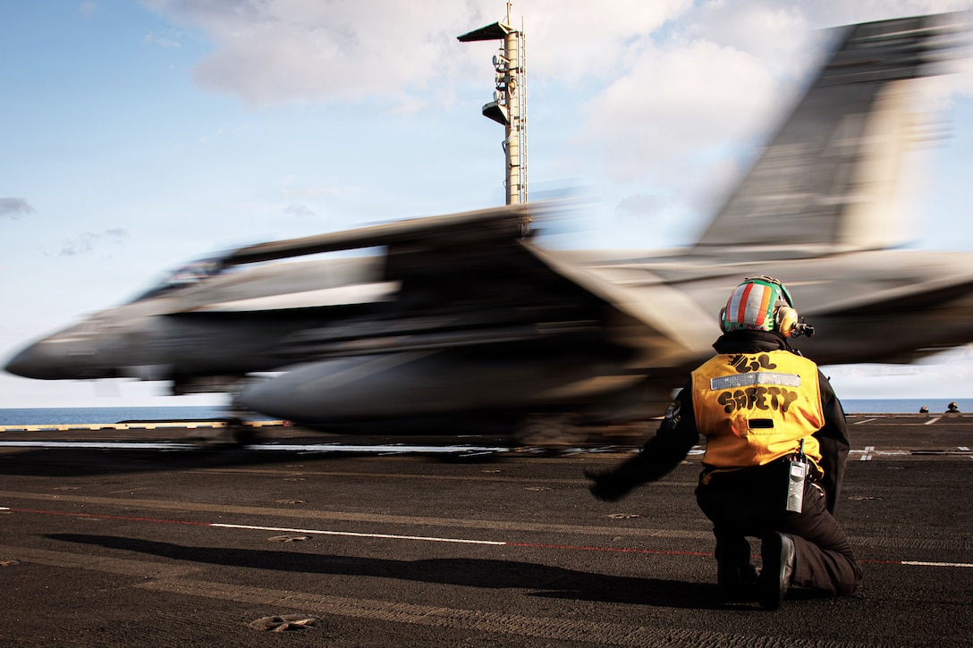 A kneeling sailor signals to a blurry aircraft as it launches from a ship at sea.