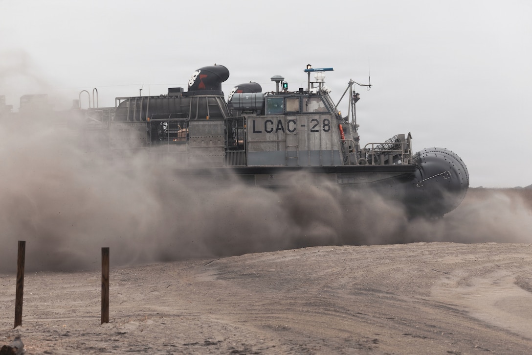 An air-cushioned landing craft drives on the sand, creating large clouds of smoke on a gloomy day.