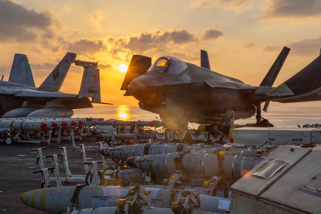 Aircraft and ordnance sit on the flight deck of a ship at sea under a sunlit sky.