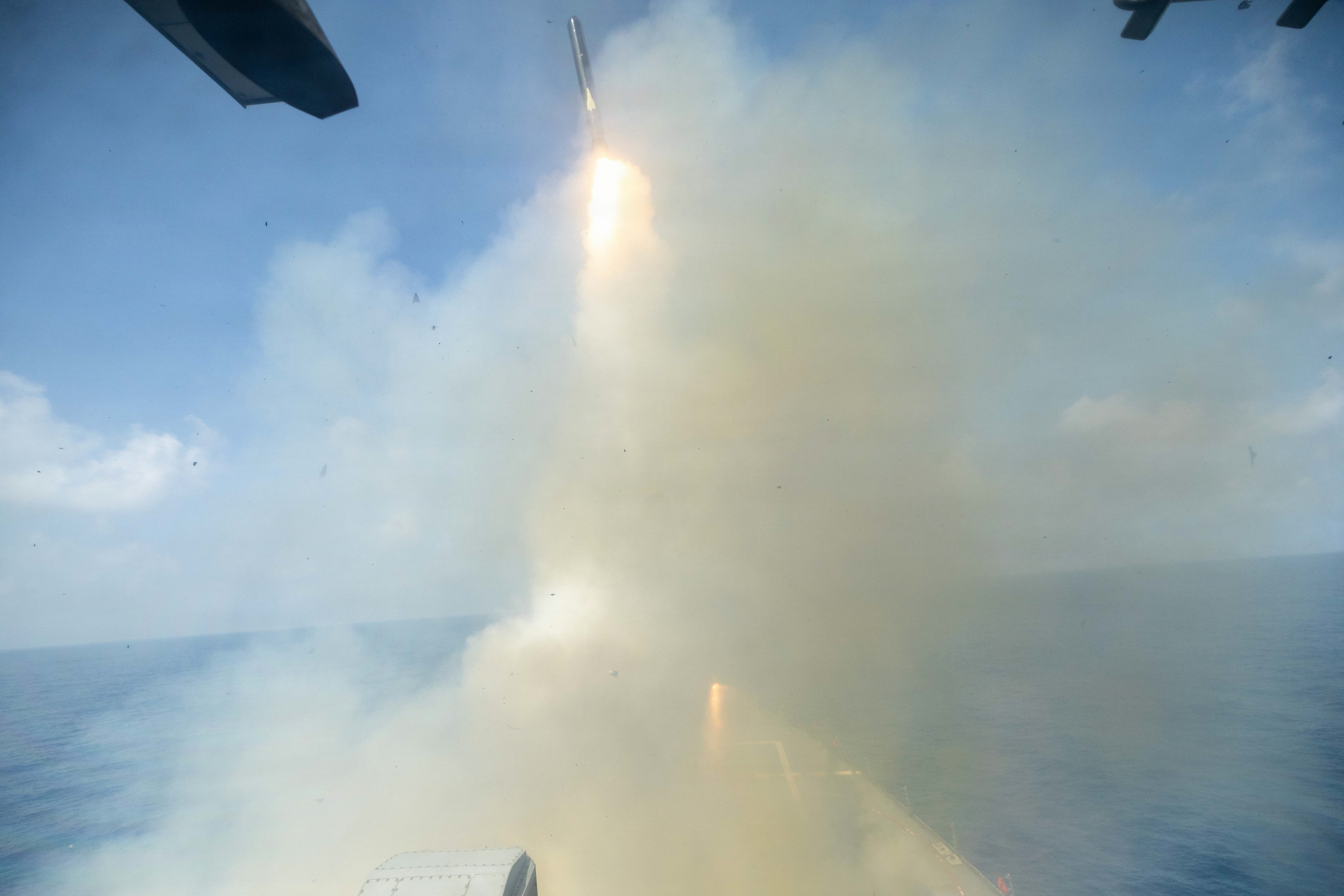 A missile launches from a ship at sea during daytime, leaving a trail of flame and billowing smoke.
