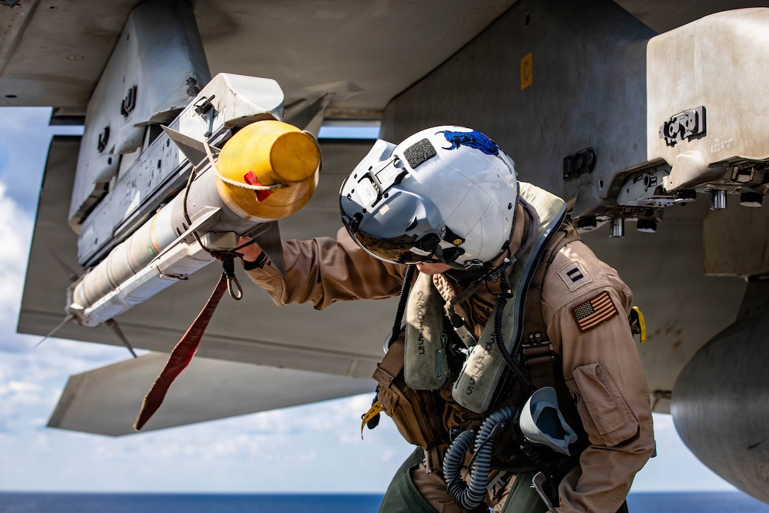A sailor looks at a yellow-tipped missile on an aircraft parked on a flight deck at sea during daytime.