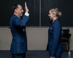 An Air Force officer administers the oath of office to a female service member. Both are in blue service dress uniforms, facing each other in profile against a dark backdrop. He has his right hand raised while she stands at attention with a slight smile.