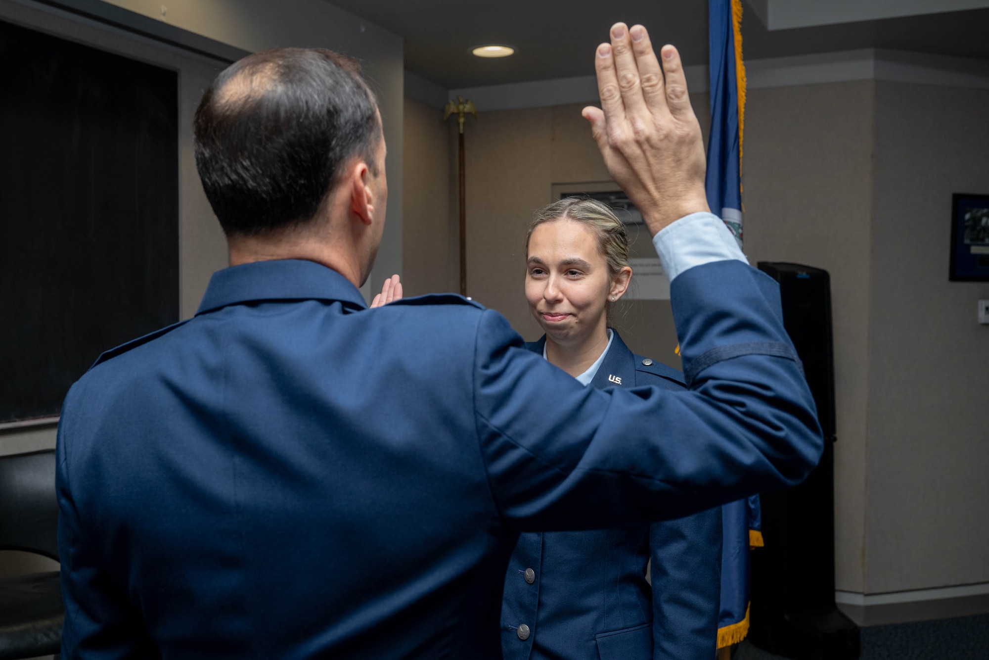 An Air Force officer, seen from behind with his right hand raised, administers the oath of office to a female Air Force member. She stands at attention, facing him, while an American flag is visible in the background.