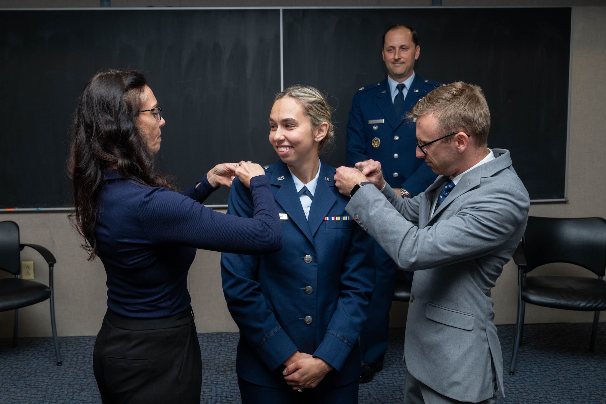 A female Air Force member in a blue service dress uniform smiles as a woman and a man in civilian attire pin new rank insignia onto her shoulders. An Air Force officer stands smiling in the background.