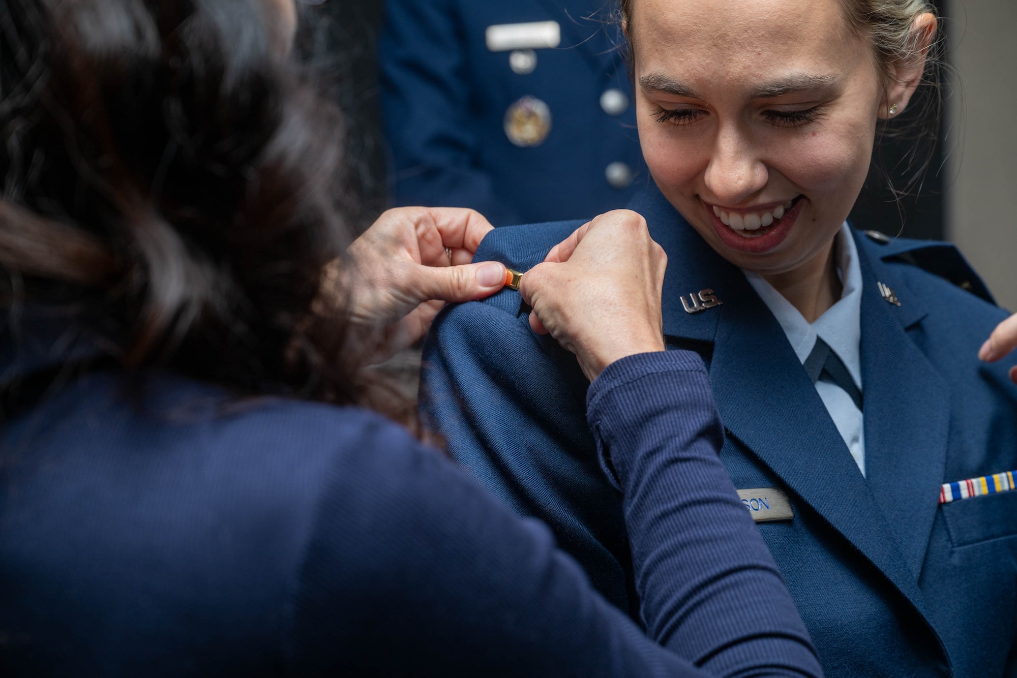 A close-up shot shows a smiling female Air Force member in a blue service dress uniform having a second lieutenant insignia pinned to her shoulder by another person. The focus is on the smiling service member and the hands carefully adjusting the rank on her epaulet.