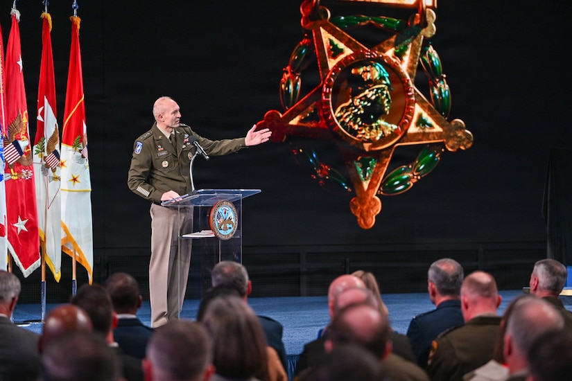 A man wearing a formal military uniform stands indoors on a stage behind a lectern, gesturing with his left hand. An image of a large military medal is on the right; four assorted flags are on the left, and in front of him is an audience.