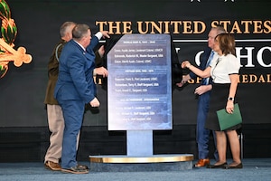 Four people, some wearing business attire and one in a formal military uniform, stand on a stage and pull a black cloth off a large plaque that lists a series of names.