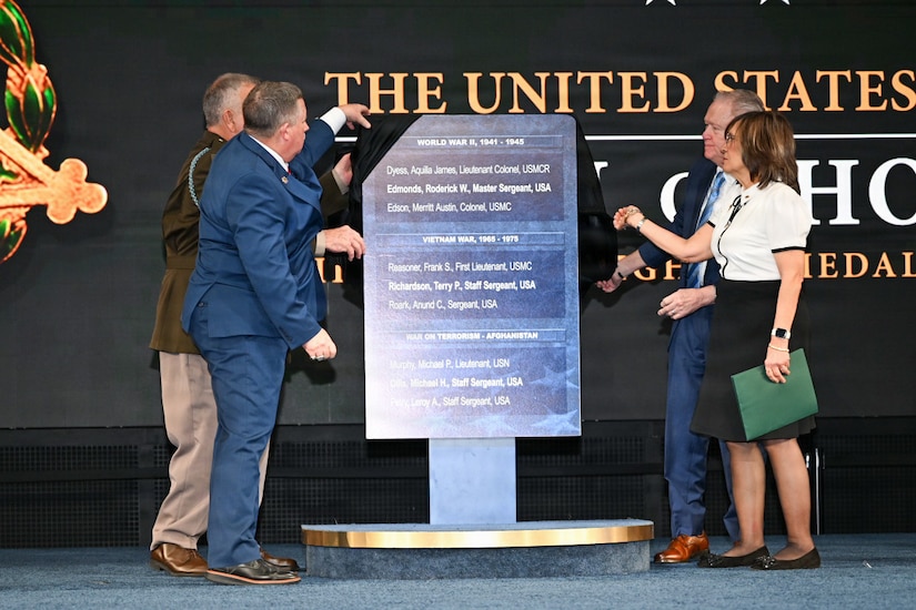 Four people, some wearing business attire and one in a formal military uniform, stand on a stage and pull a black cloth off a large plaque that lists a series of names.