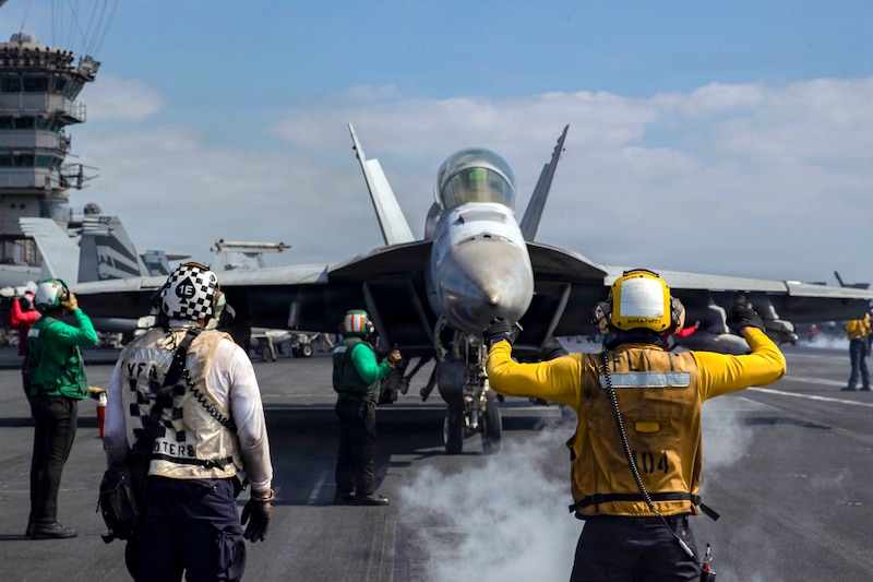 A sailor in a yellow shirt and helmet signals to an aircraft on a ship's flight deck, with other sailors working nearby.