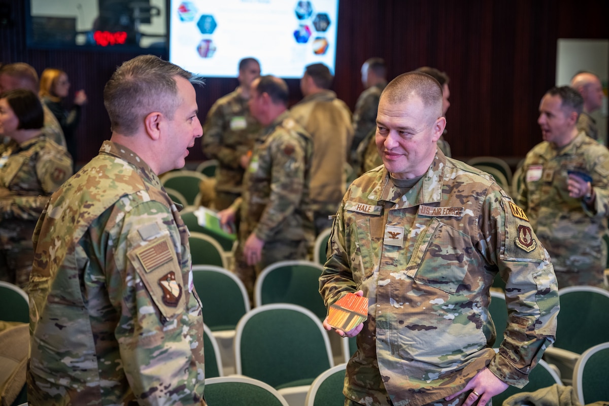Two men, wearing camouflage military uniforms, stand while talking to each other. Other military members can be seen in background