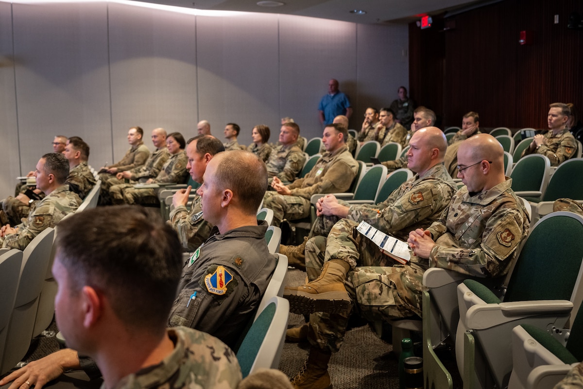 Military members, in camouflage uniforms, fill a small auditorium, some taking notes