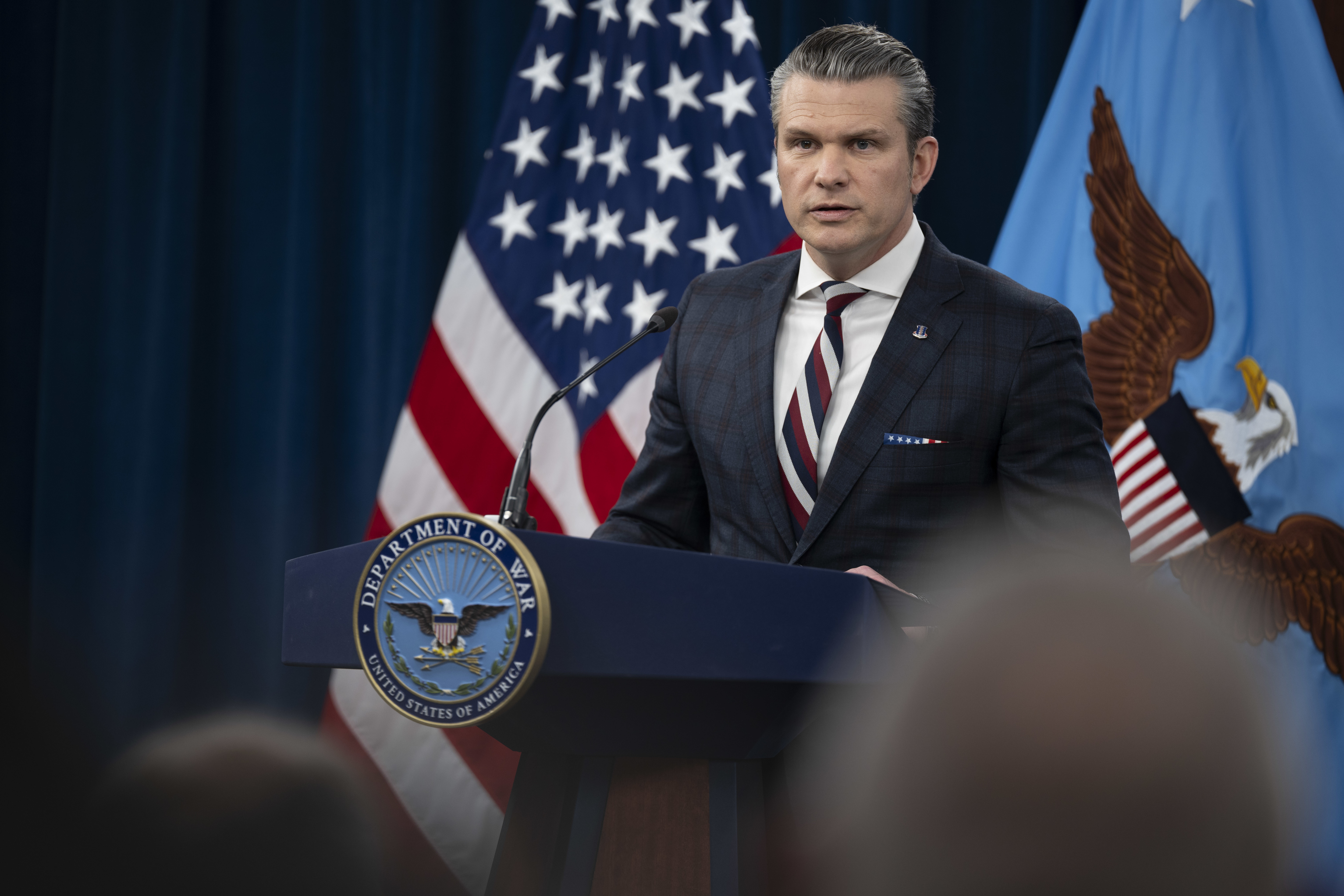 A man in business attire stands behind a lectern and speaks; behind him are an American flag and a blue flag with an eagle in the center.