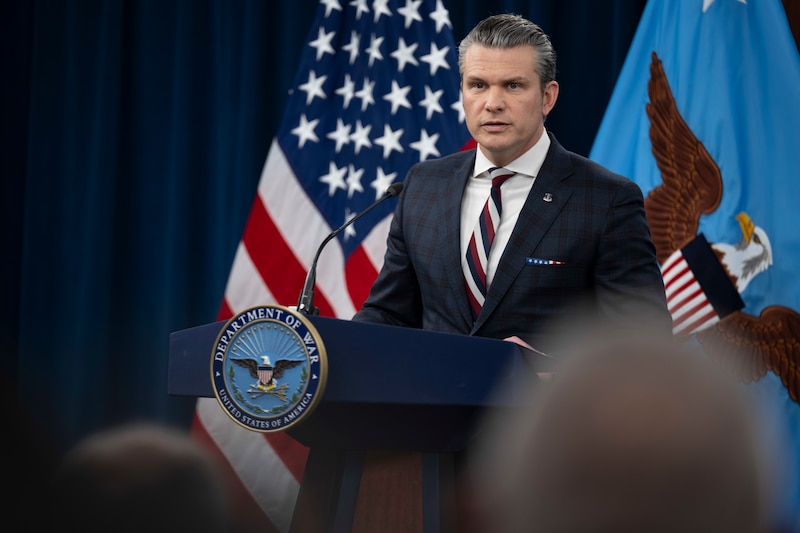 A man in business attire stands behind a lectern and speaks; behind him are an American flag and a blue flag with an eagle in the center.