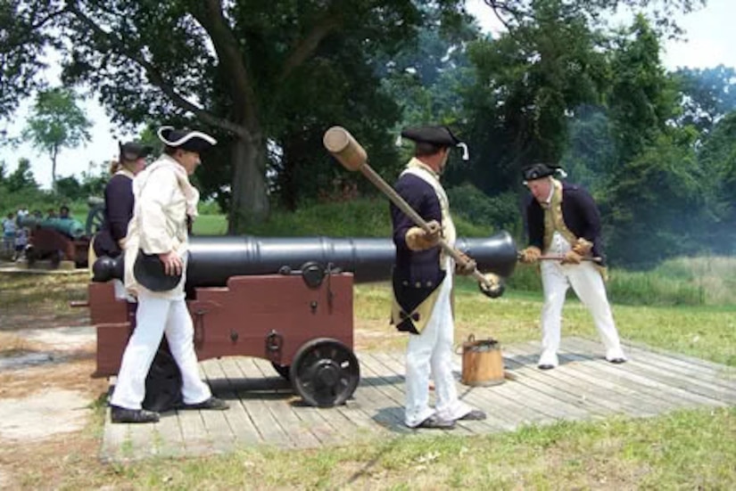 Men in colonial military uniforms prepare to fire a cannon in a field. There are trees in the background.