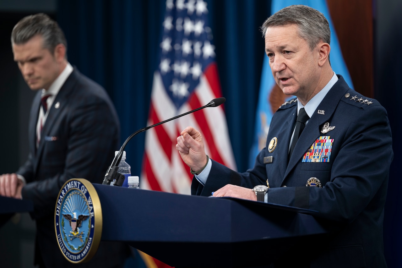 A man in a formal military uniform and a man in business attire stand at lecterns and speak, with an American flag behind them.