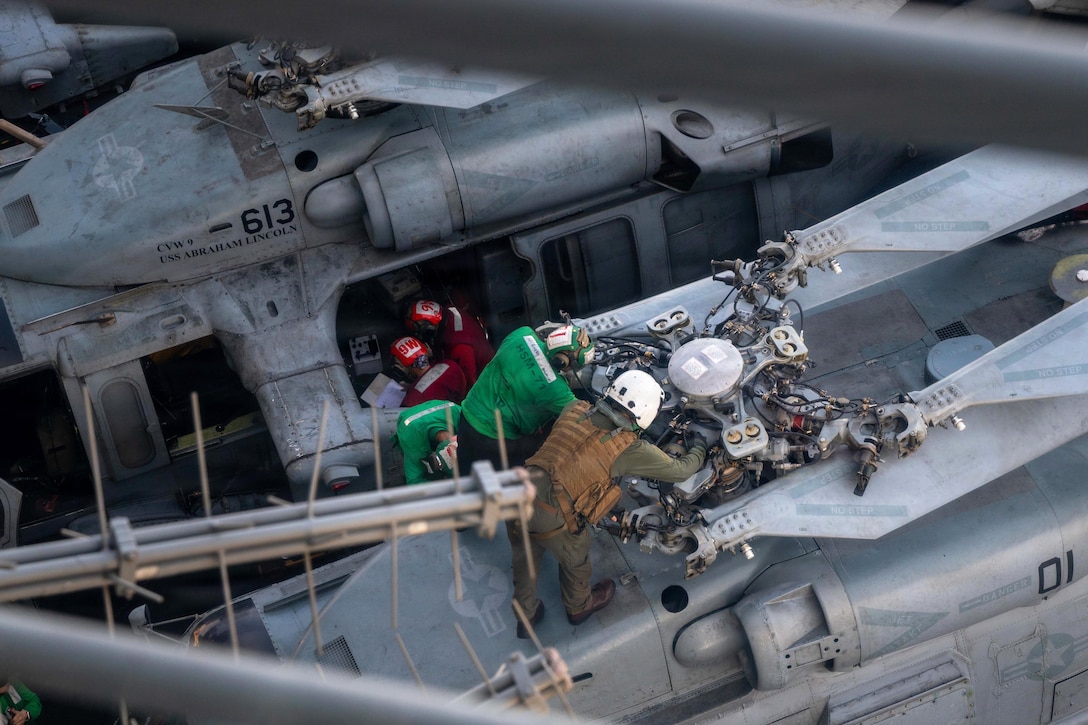 Sailors in protective gear work on a helicopter aboard a ship as seen from above.