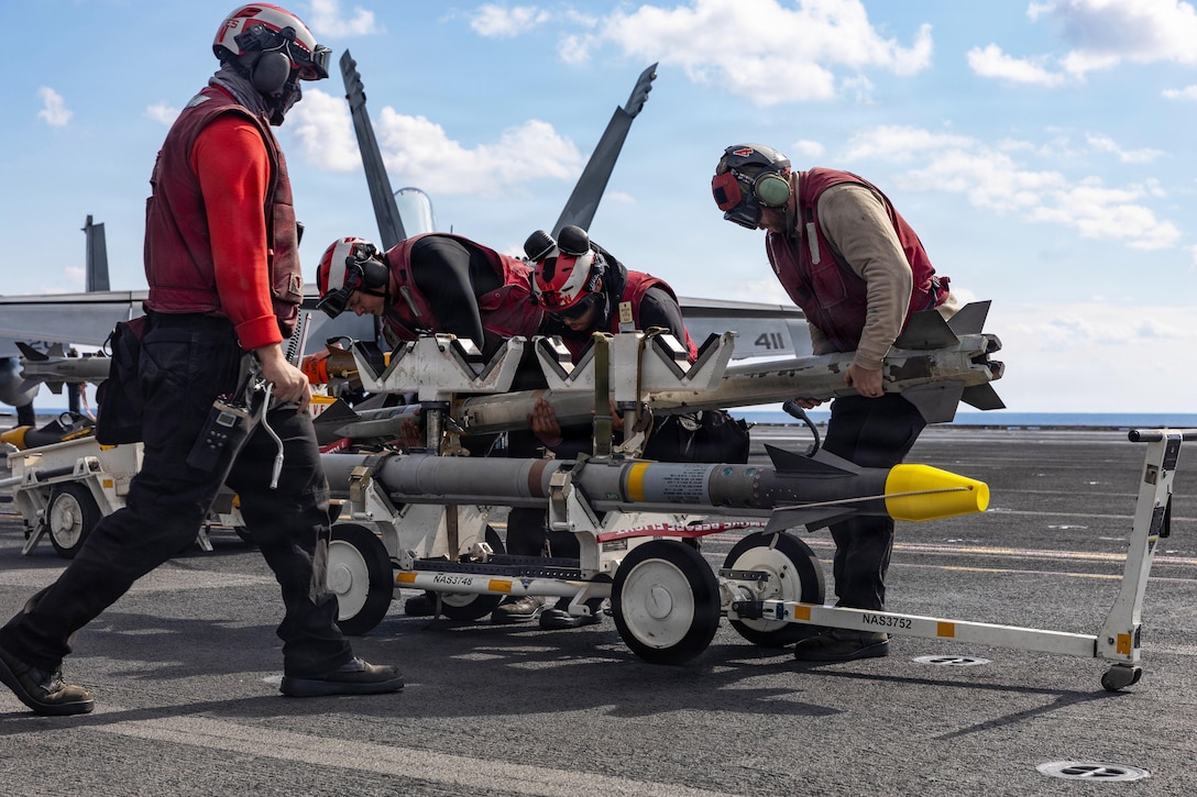 Four sailors in red helmets and vests handle ordnance aboard a ship at sea during the day.