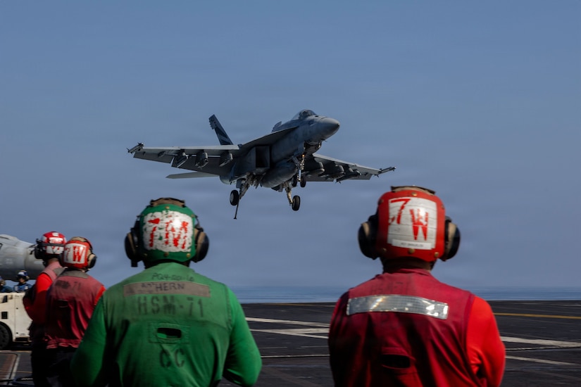 A military aircraft lands on an aircraft carrier flight deck as four people wearing protective helmets look on from the foreground.