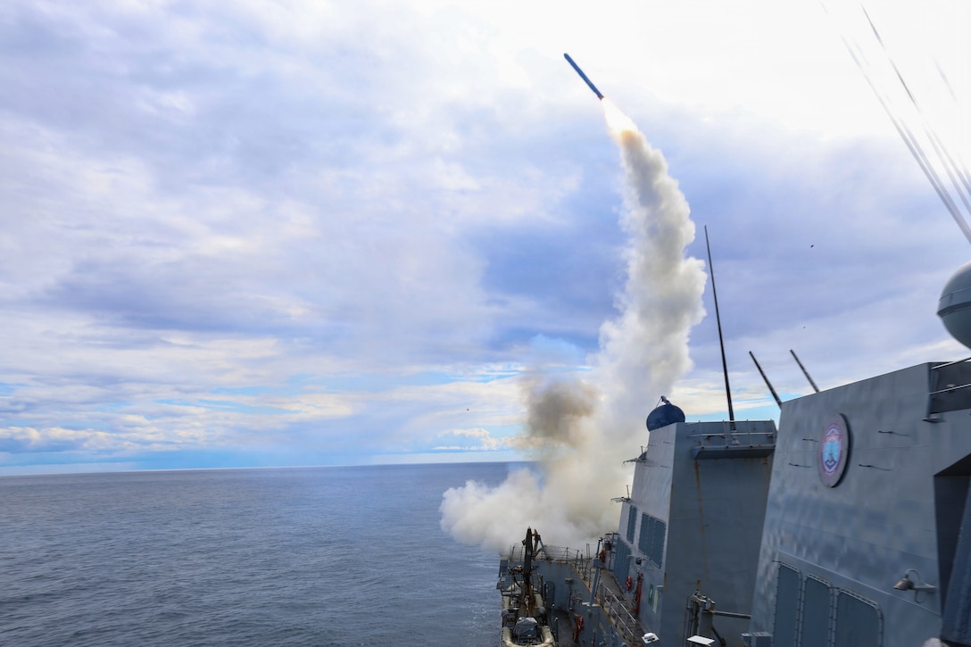 A missile launches from a ship at sea during the day, leaving behind a trail of smoke.