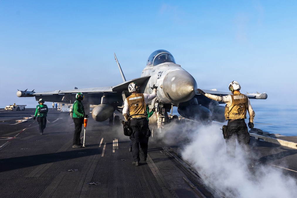 Sailors stand on a smoky flight deck around an aircraft under a blue sky.
