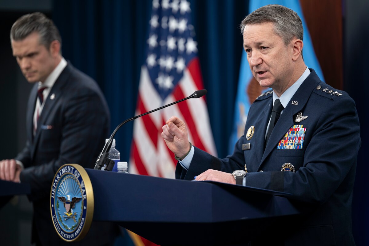 A man in a formal military uniform and a man in business attire stand at lecterns and speak, with an American flag behind them.