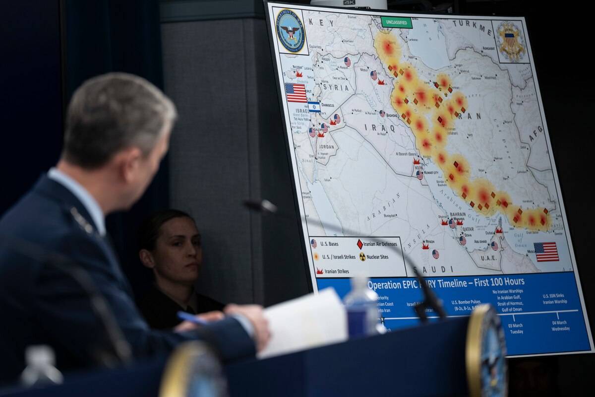 A man in a formal military uniform stands at a lectern, looking back at a map of the Middle East with symbols; another person looks on in the background.