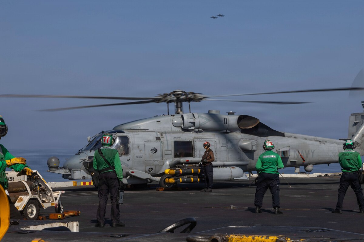 A military helicopter is parked on the flight deck of a ship. A crew member walks on the deck near the helicopter, with the ocean and a hazy sky in the background. Four people wearing protective helmets are looking at the helicopter from the foreground.