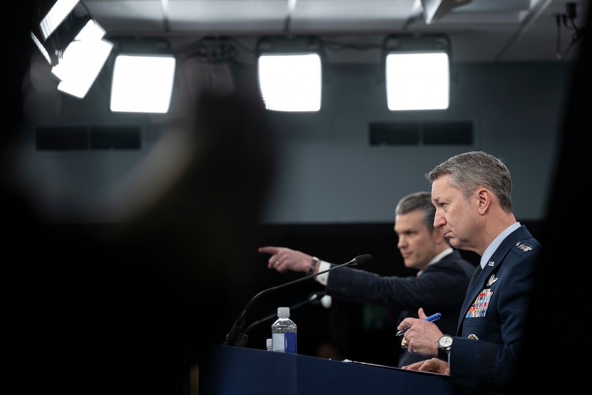 A man in a business suit and a man in a formal military uniform stand at lecterns; the man in a suit points forward with one hand.