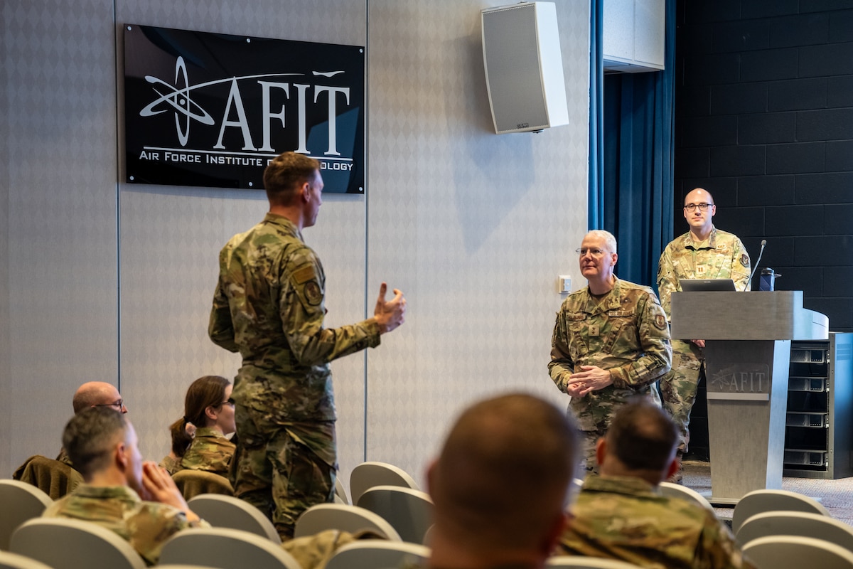 Two men, wearing camouflage military uniforms, stand in a small auditorium addressing each other as other members are seated