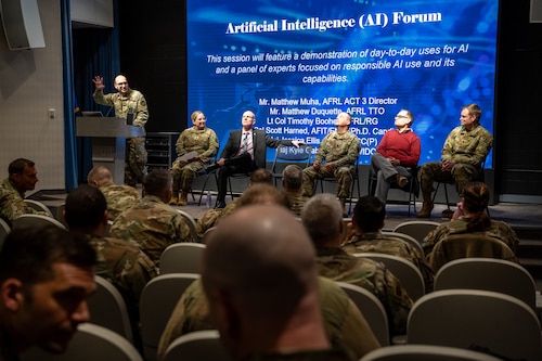 Five people can be seen sitting on a stage, one standing at a podium, in front of a blue screen with names.
