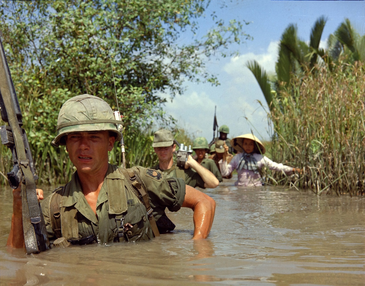 A man carrying an automatic rifle wades into chest deep swamp water. At least four people follow behind him.