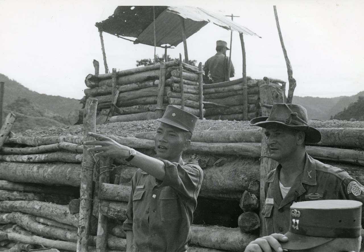 Two men look into the distance as one points. Behind them is a log fence and a taller lookout post also made of logs.